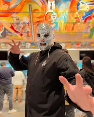 Person in a metallic Lucha Libre mask posing dramatically in front of a colorful, action-packed wrestling-themed mural.