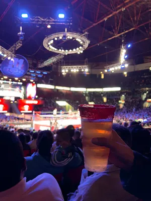 A chili-rimmed craft michelada held up at a Lucha Libre wrestling match inside a bright, energetic sports arena.