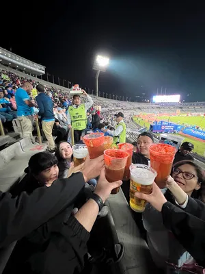 A group of fans raises a craft michelada in a stadium toast during a night soccer match under the bright lights.