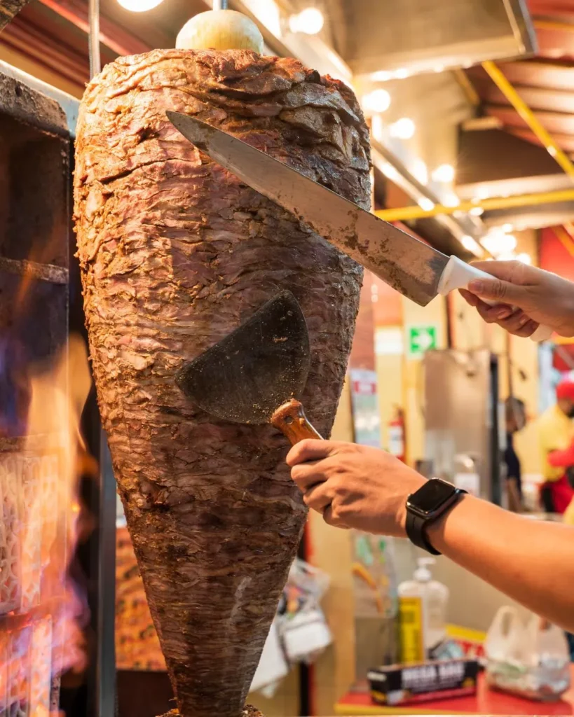 Close up of a chef slicing marinated pork from a vertical spit to prepare different types of tacos al pastor.