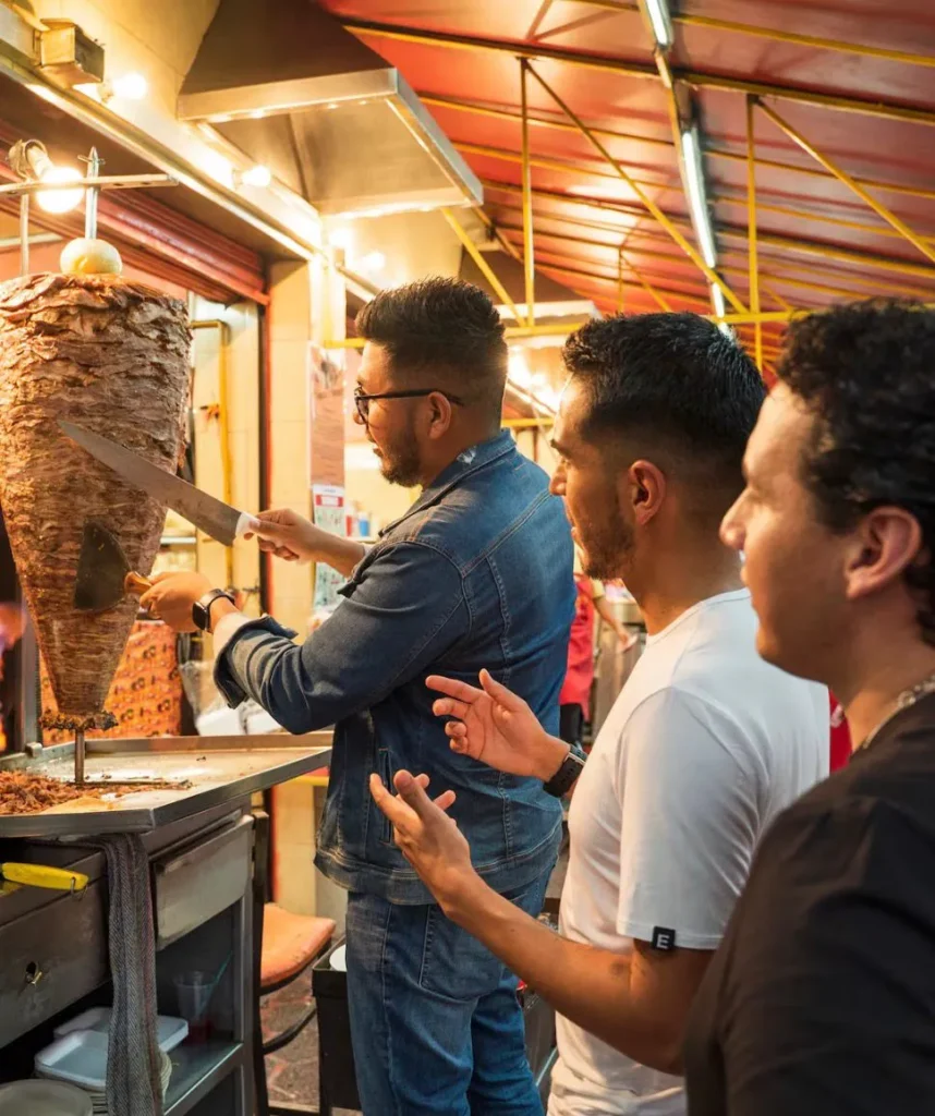 Three friends watching a taquero slice meat from a traditional pastor trompo for the best tacos in Mexico City.