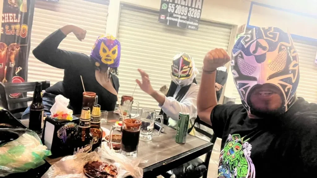 Three people in colorful wrestling masks enjoy drinks at a bar while discovering what is a michelada in Mexico City.