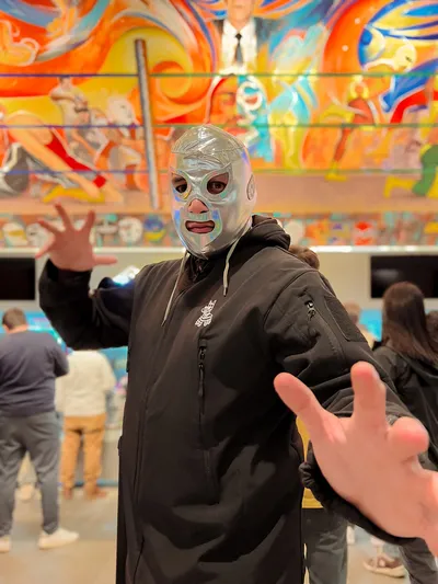 A man wearing a shiny silver wrestling mask poses in front of a colorful mural at Lucha Libre in Mexico City.