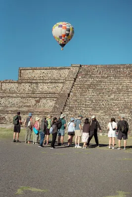 A group of travelers standing near ancient ruins watching a colorful sunrise hot air balloon ride over Teotihuacán.